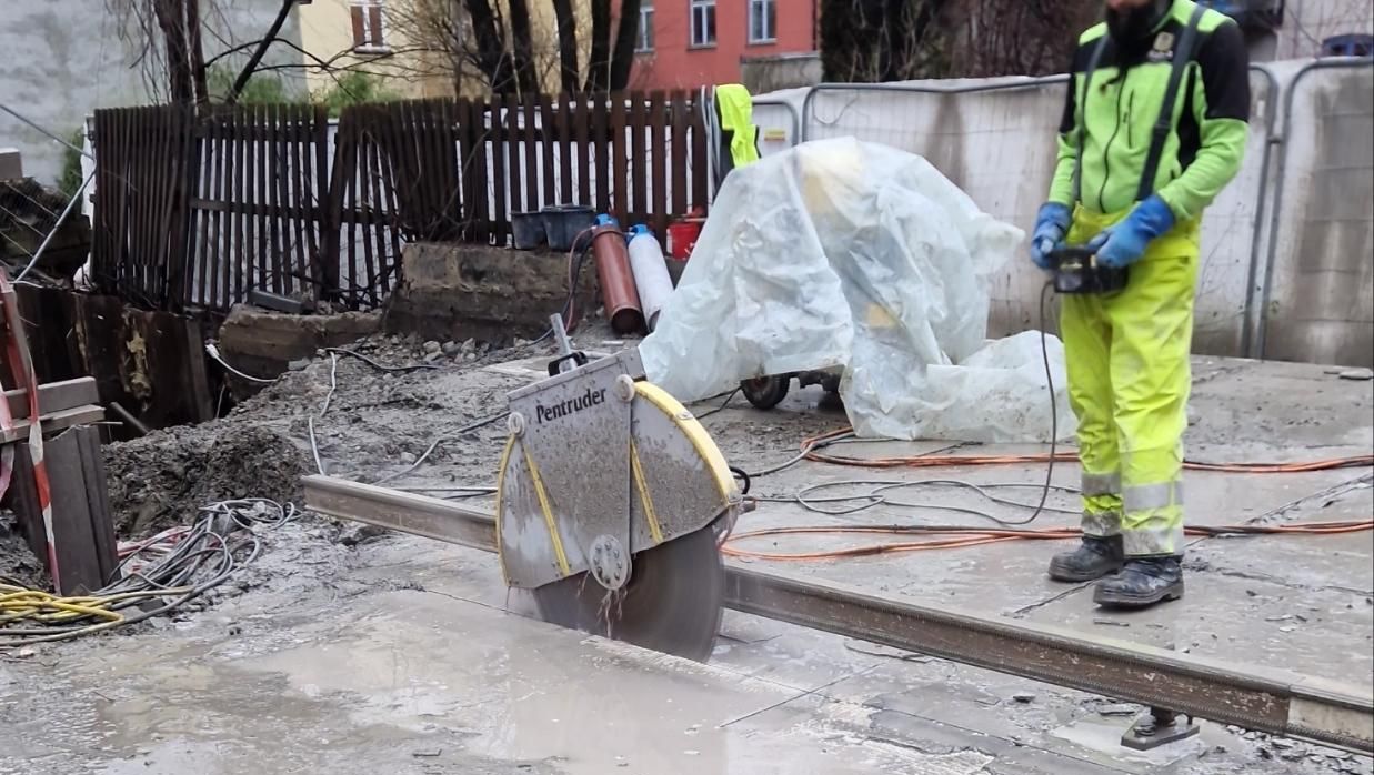 En arbeider betjener en stor betongsag på skinner som skjærer gjennom våt betong på byggeplass.