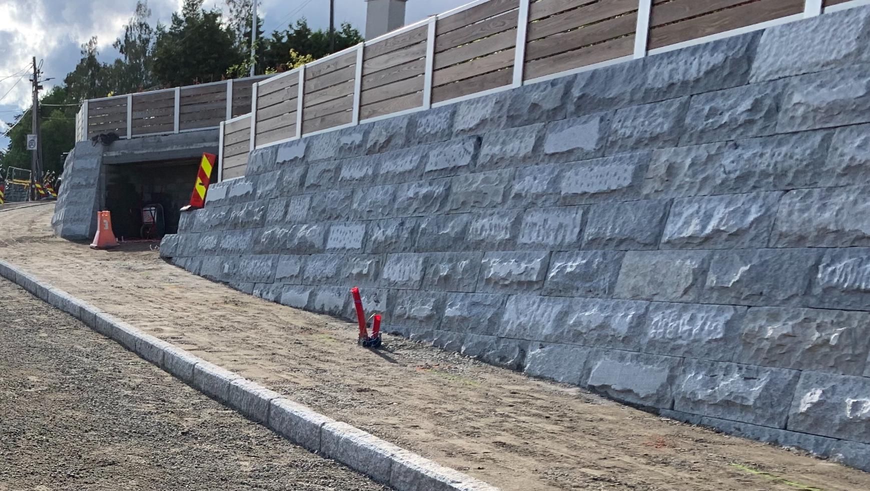 Ny støttemur av steinblokker langs gangvei med rekkverk på toppen og noen markerte kabler.